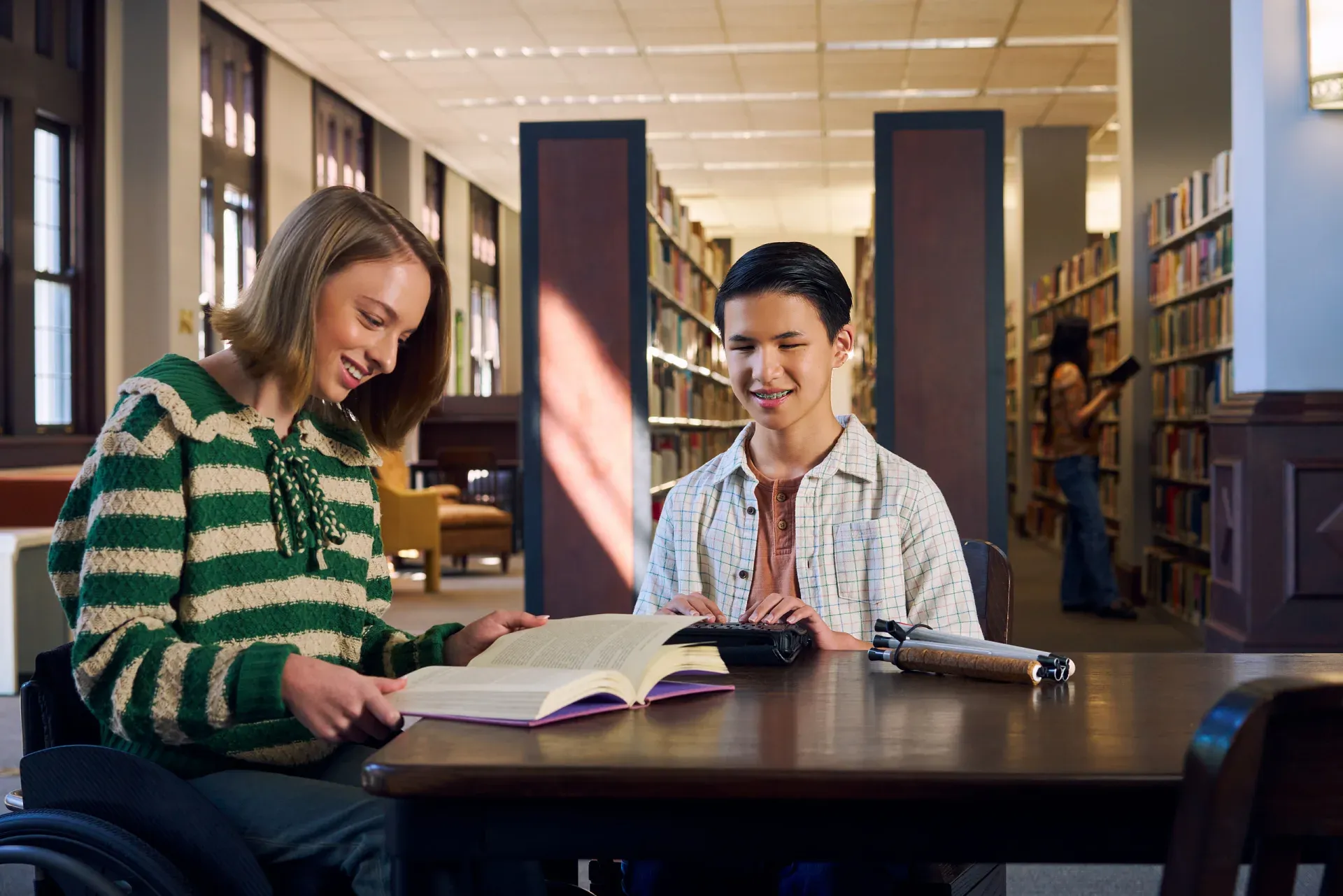 Two students reading together at a library table with mobility aids nearby.